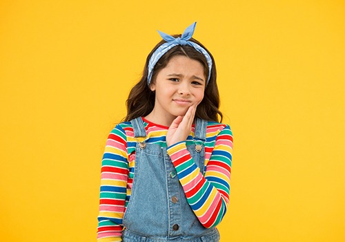 Girl in striped shirt and overalls touching jaw in pain