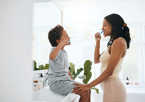 Little boy sitting on bathroom counter brushing teeth facing a woman brushing teeth