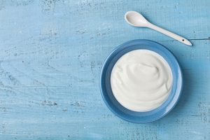 Bird's eye view of plain yogurt in blue bowl on wood surface next to small spoon